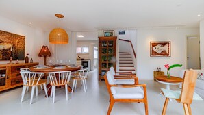 A spacious living area with a dining table, white chairs, a wooden coffee table, and a staircase leading to another level. - MSP01 Apt 3 Quartos Pé na Areia (Cairu)