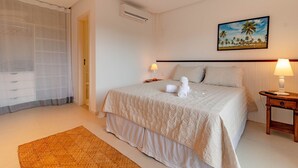 A neatly made bed with a white duvet, a small bedside table with a lamp, and a painting of a tropical scene on the wall. - MSP01 Apt 3 Quartos Pé na Areia (Cairu)