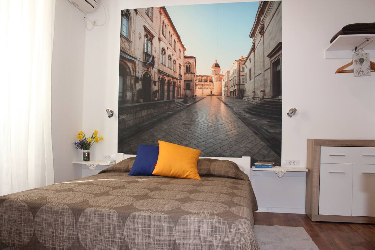 Ein Hotelzimmer mit einem großen Bett, einem Nachttisch mit Blumen in einer Vase und Blick auf eine belebte Stadtstraße durch ein großes Fenster.