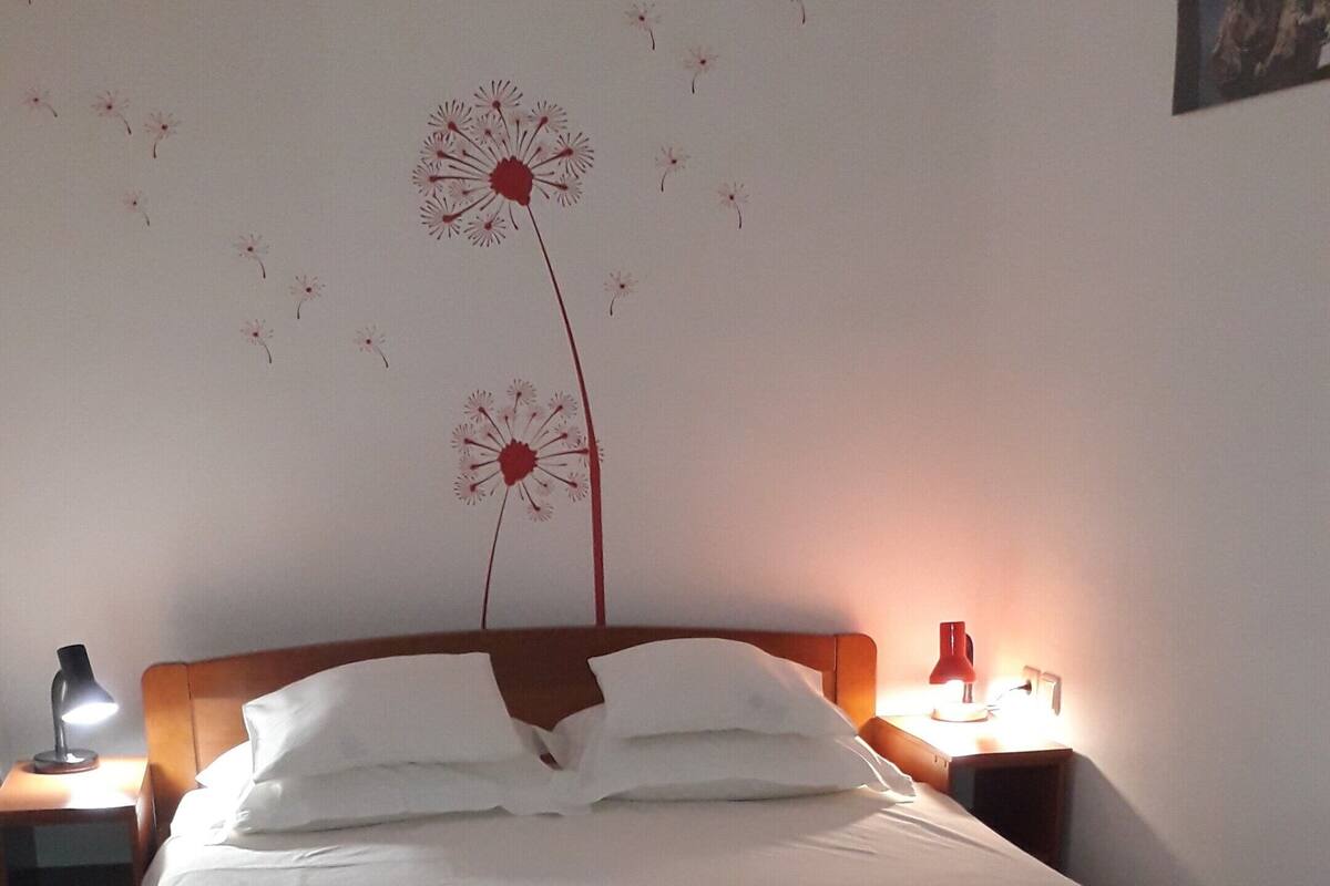 Ein ordentlich bezogenes Bett mit weißen Leinentüchern, ein hölzernes Kopfteil und eine gelbe Bank am Fußende. Zwei Nachttische mit Lampen, und eine Wanddekoration mit Löwenzahn.