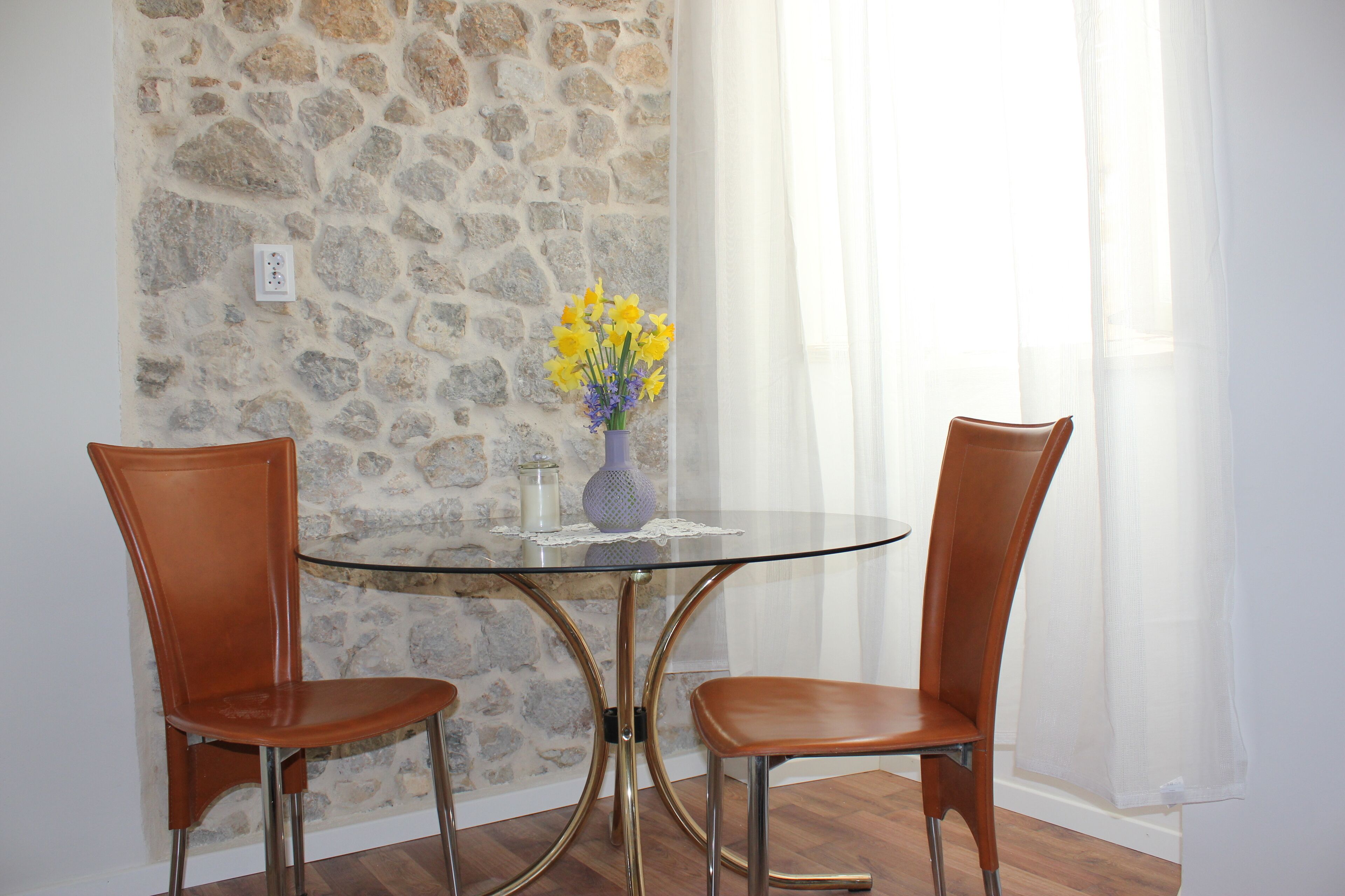 Un comedor con una mesa de vidrio rodeada por dos sillas marrones, un jarrón con flores amarillas y una pared de piedra.