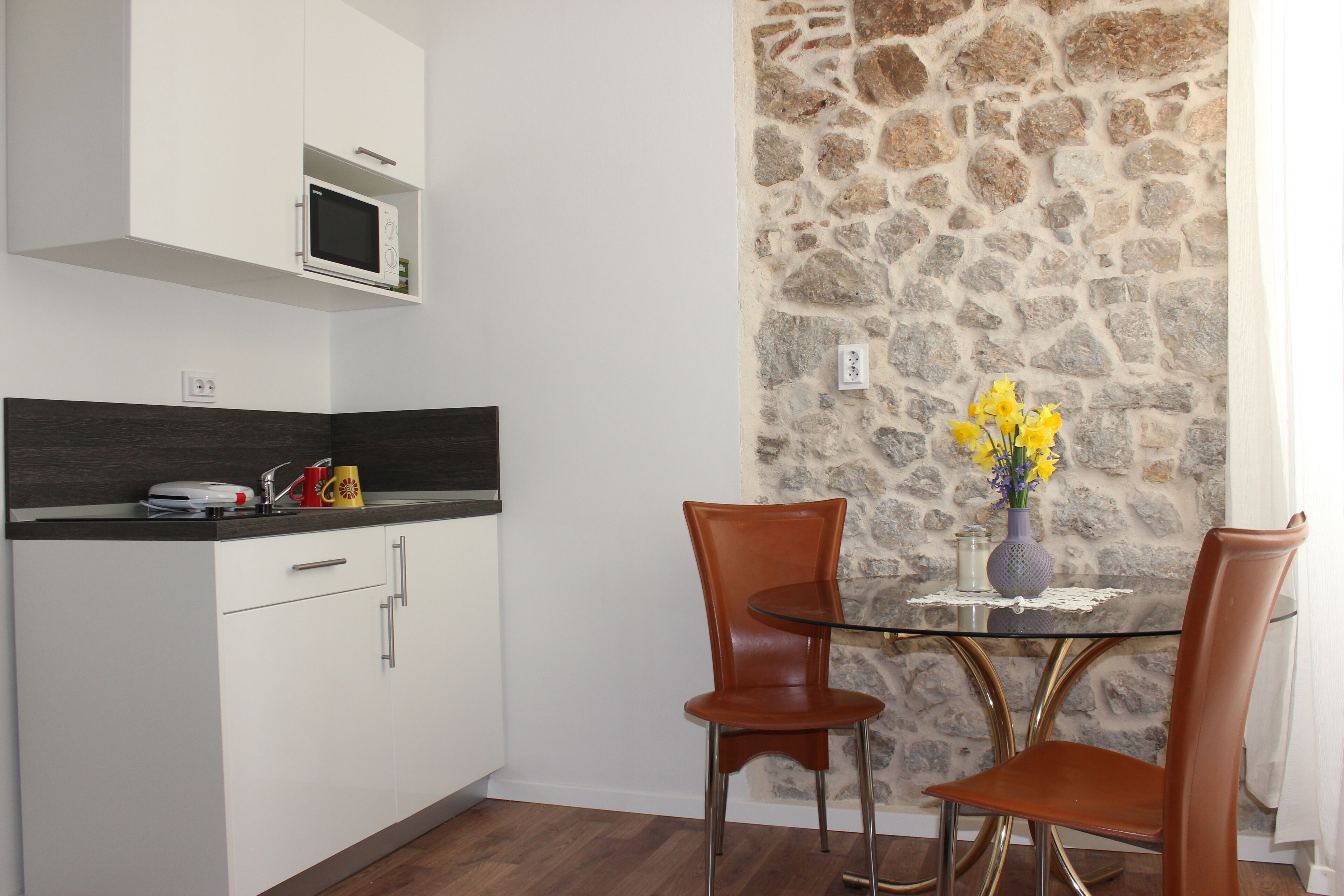 A compact kitchen with white cabinets, a microwave, and a small table with two chairs. A stone wall and a vase with flowers are visible in the background.