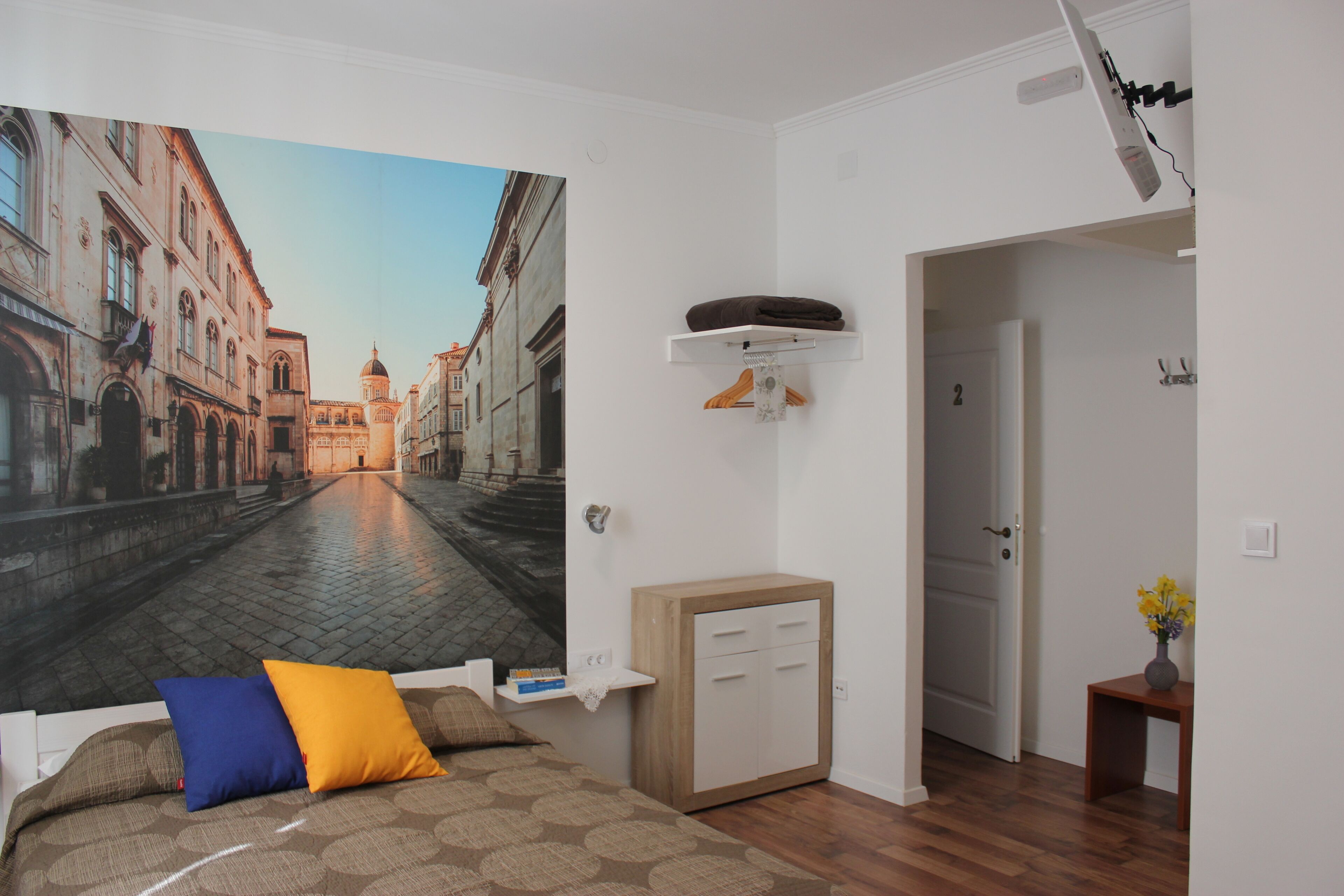 Una habitación con un amplio mural de la ciudad en la pared, una cama con ropa de cama estampada, un pequeño armario de madera y una puerta que da a otra habitación.