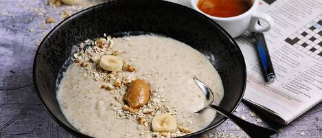 A bowl of oatmeal with banana slices and nuts, a spoon, and a cup of tea on a table with a newspaper.