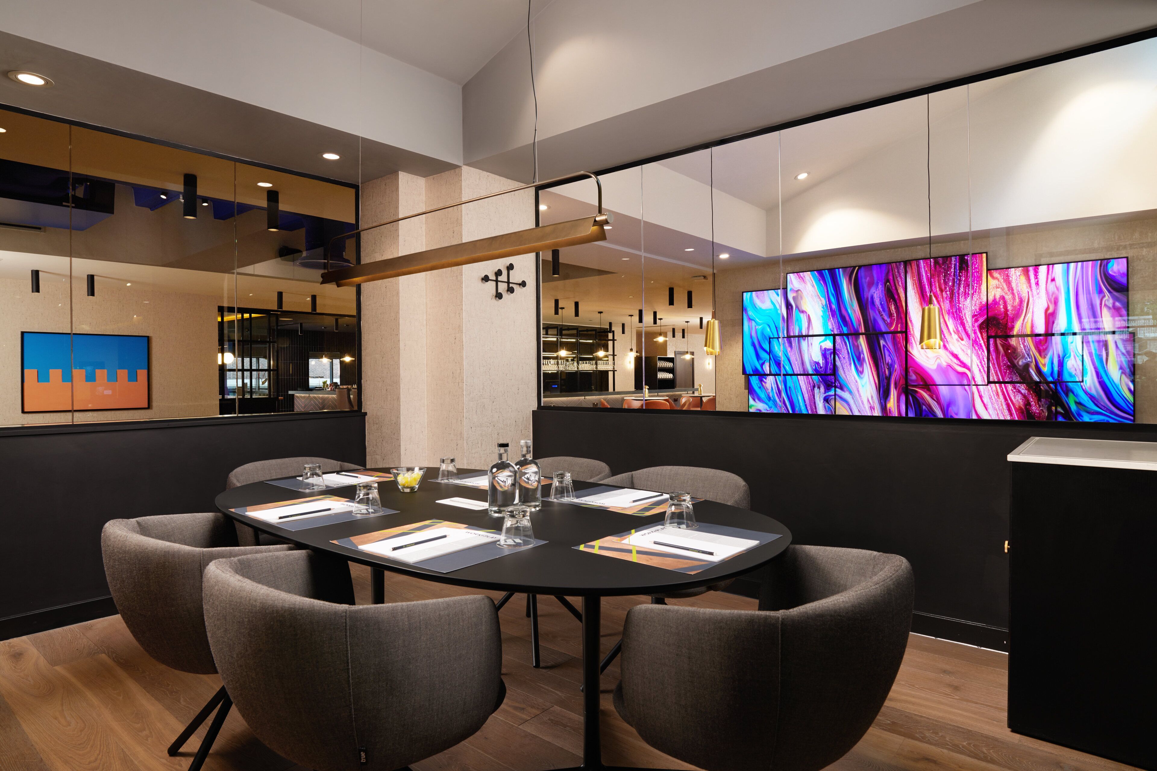 A modern conference room with a round table, grey upholstered chairs, and a large abstract painting on the wall.