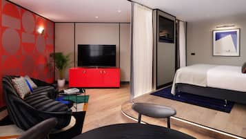 A modern hotel room with a flat-screen TV, a bed, a sofa, a red cabinet, and a potted plant.