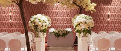A wedding ceremony setup with white chairs, a floral arrangement on the altar, and a geometric patterned wall.