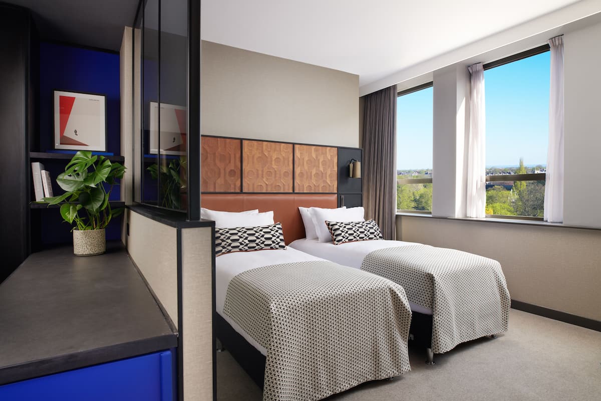Habitación de hotel con dos camas, un ventanal con vistas a la ciudad, una planta sobre una mesita de noche y una pared de acento azul.