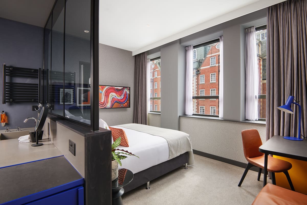Una habitación de hotel moderna con cama, escritorio, silla, baño y ventanas amplias con vistas a edificios.