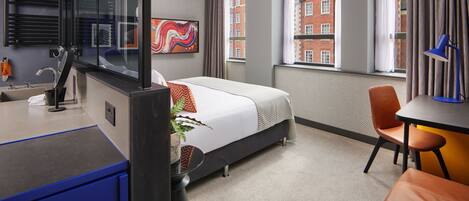 Une chambre d’hôtel moderne équipée d’un lit, d’un bureau, d’une chaise, d’une salle de bain et de grandes fenêtres donnant sur des immeubles.