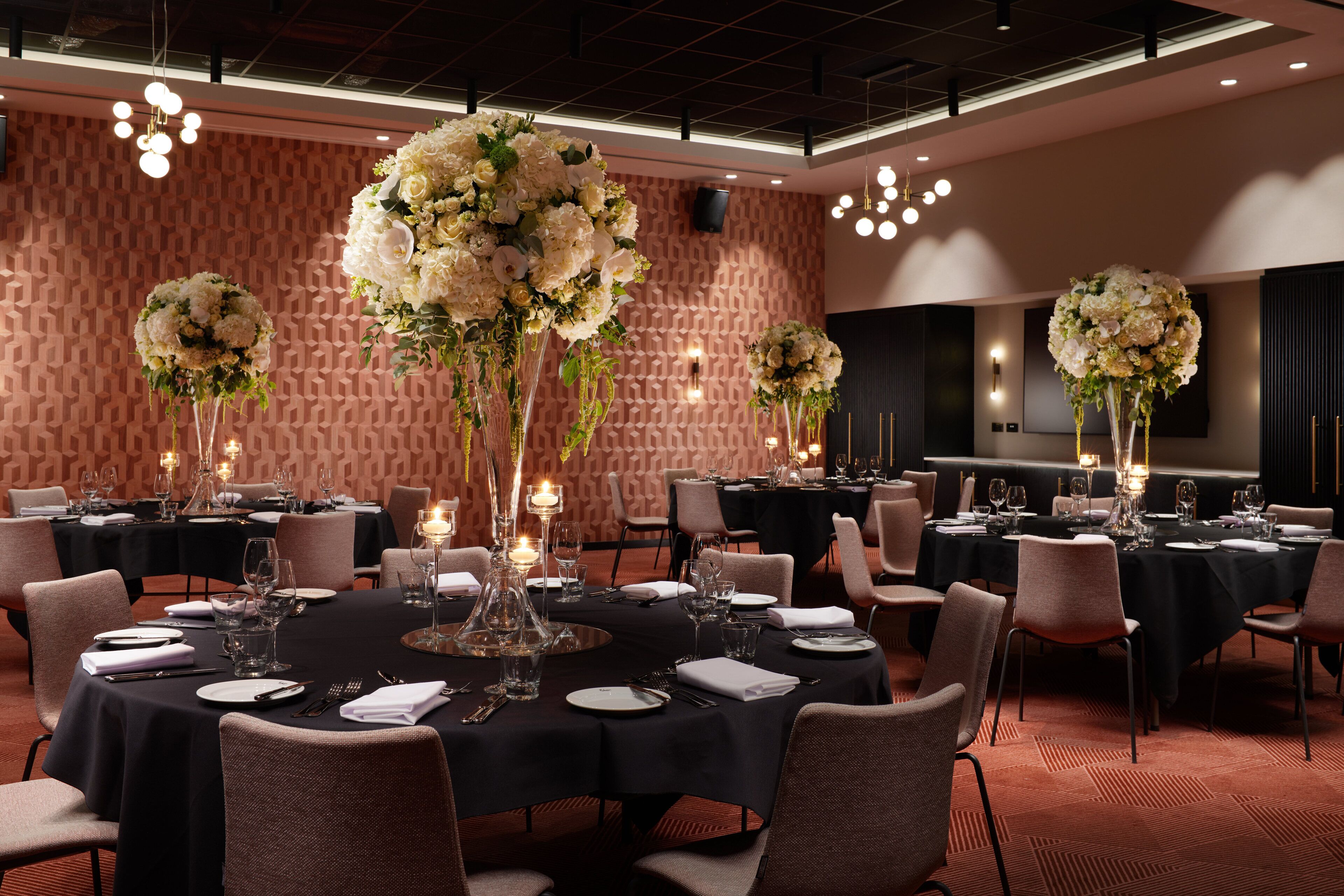 A formal dining room with round tables set for a meal, featuring floral centerpieces and candles.