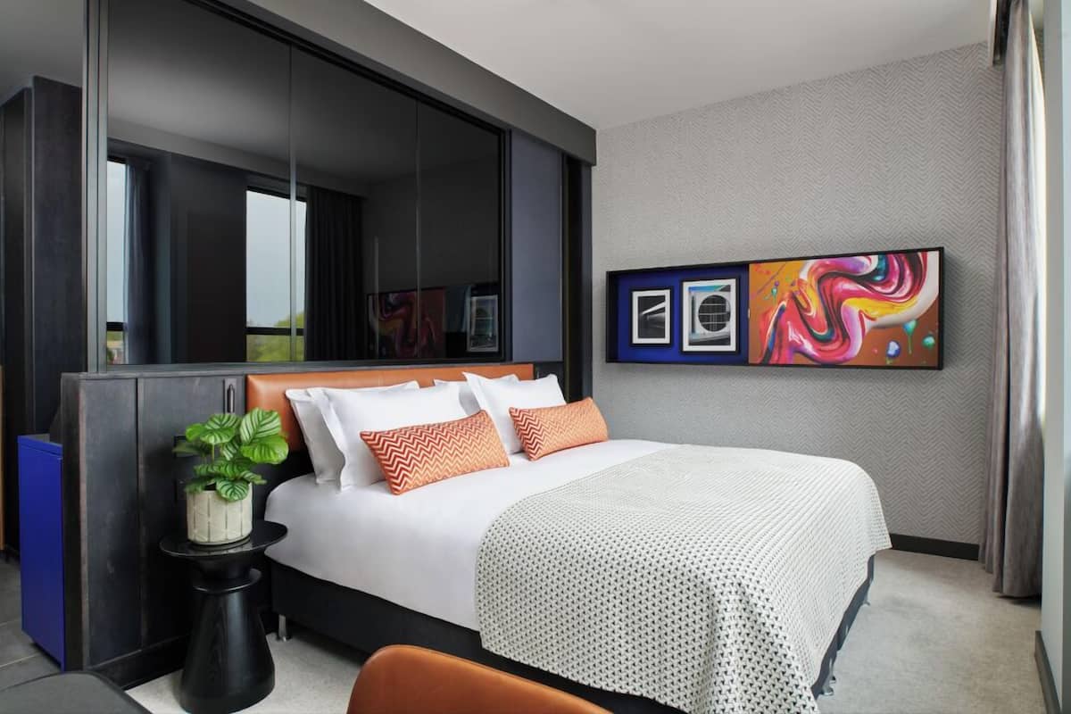 Una habitación de hotel moderna con una cama grande, una mesita auxiliar con una planta, un ventanal amplio y arte abstracto en la pared.
