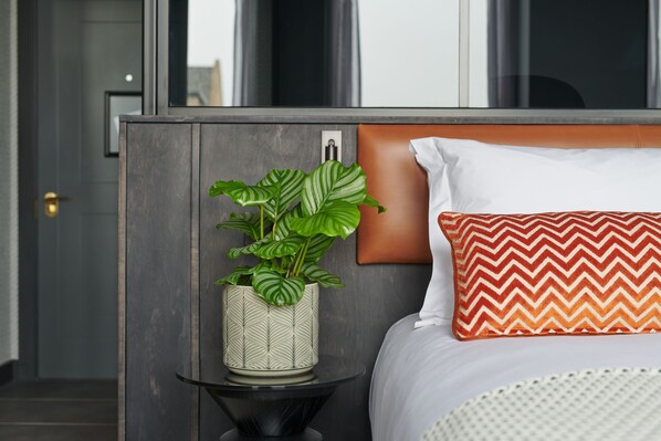 A modern hotel room with a bed, a nightstand, a potted plant, and a view of the city through large windows.