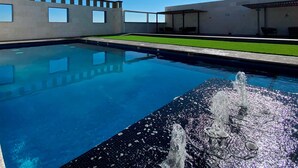 A swimming pool with a fountain feature, surrounded by a modern building with large windows and a grassy area. - Hotel MX más San Miguel de Allende (San Miguel de Allende)