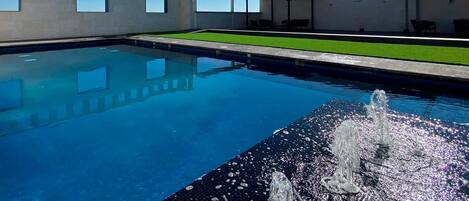 A swimming pool with a fountain feature, surrounded by a modern building with large windows and a grassy area.