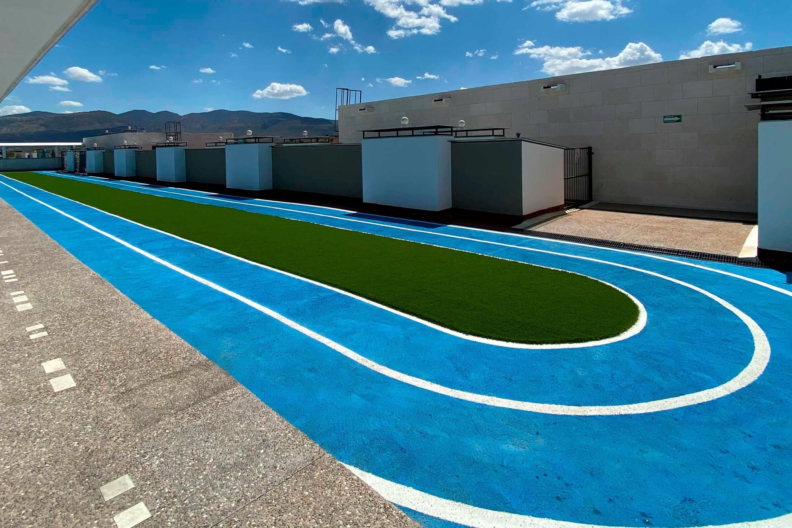 A long, straight indoor track with blue and green markings, surrounded by a concrete wall and a building with a flat roof.