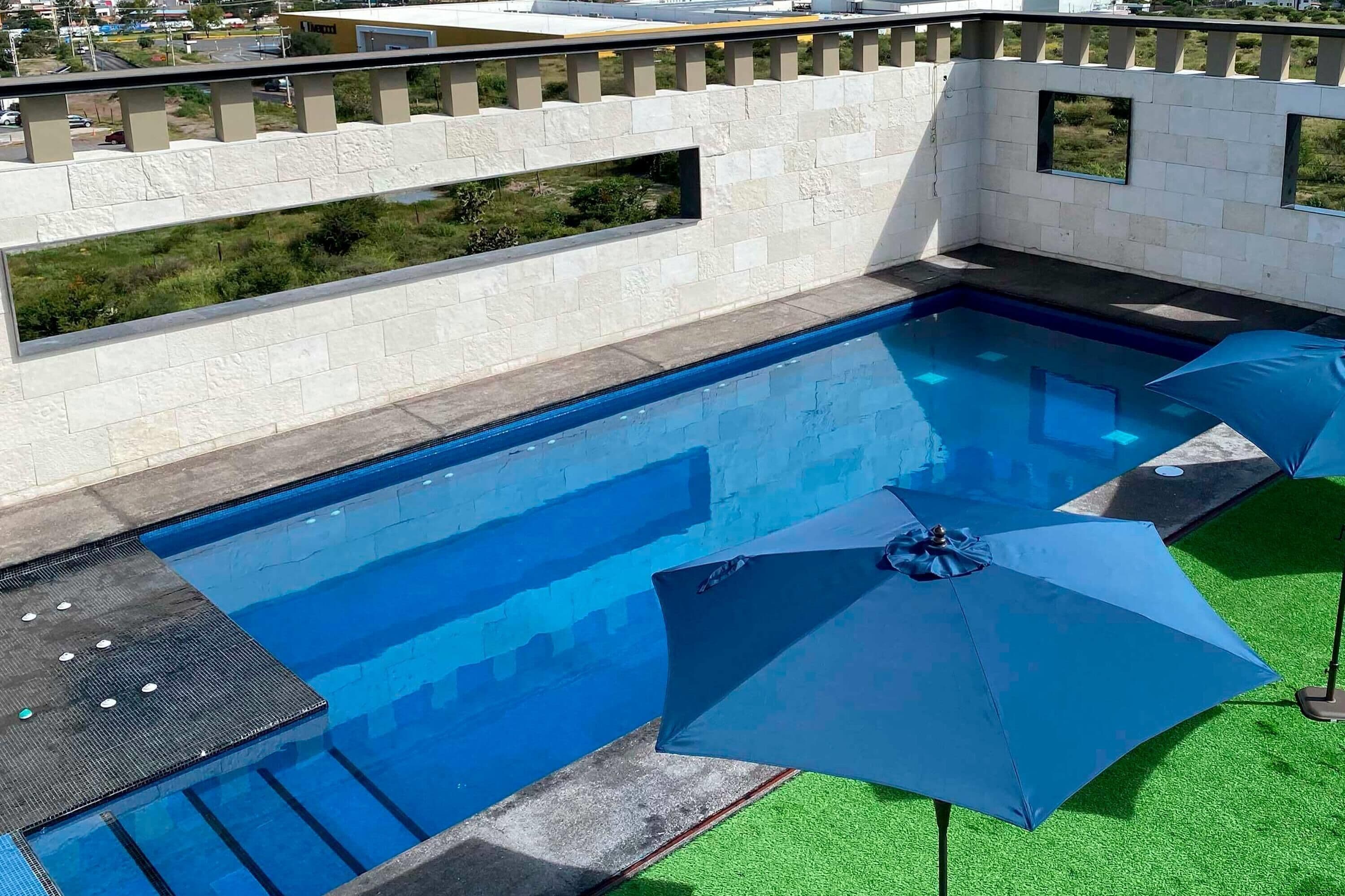 Une piscine avec un mur en pierre et des fenêtres, entourée de gazon synthétique vert et d’un parasol bleu.