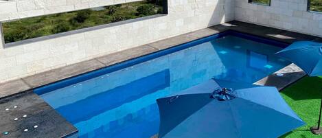 Une piscine avec un mur en pierre et des fenêtres, entourée de gazon synthétique vert et d’un parasol bleu.