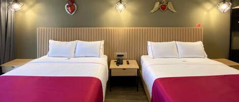 A hotel room with two beds, each with white bedding and red throw pillows. There are bedside tables with lamps, a phone, and a small table between the beds. The walls have decorative heart and angel wall art.