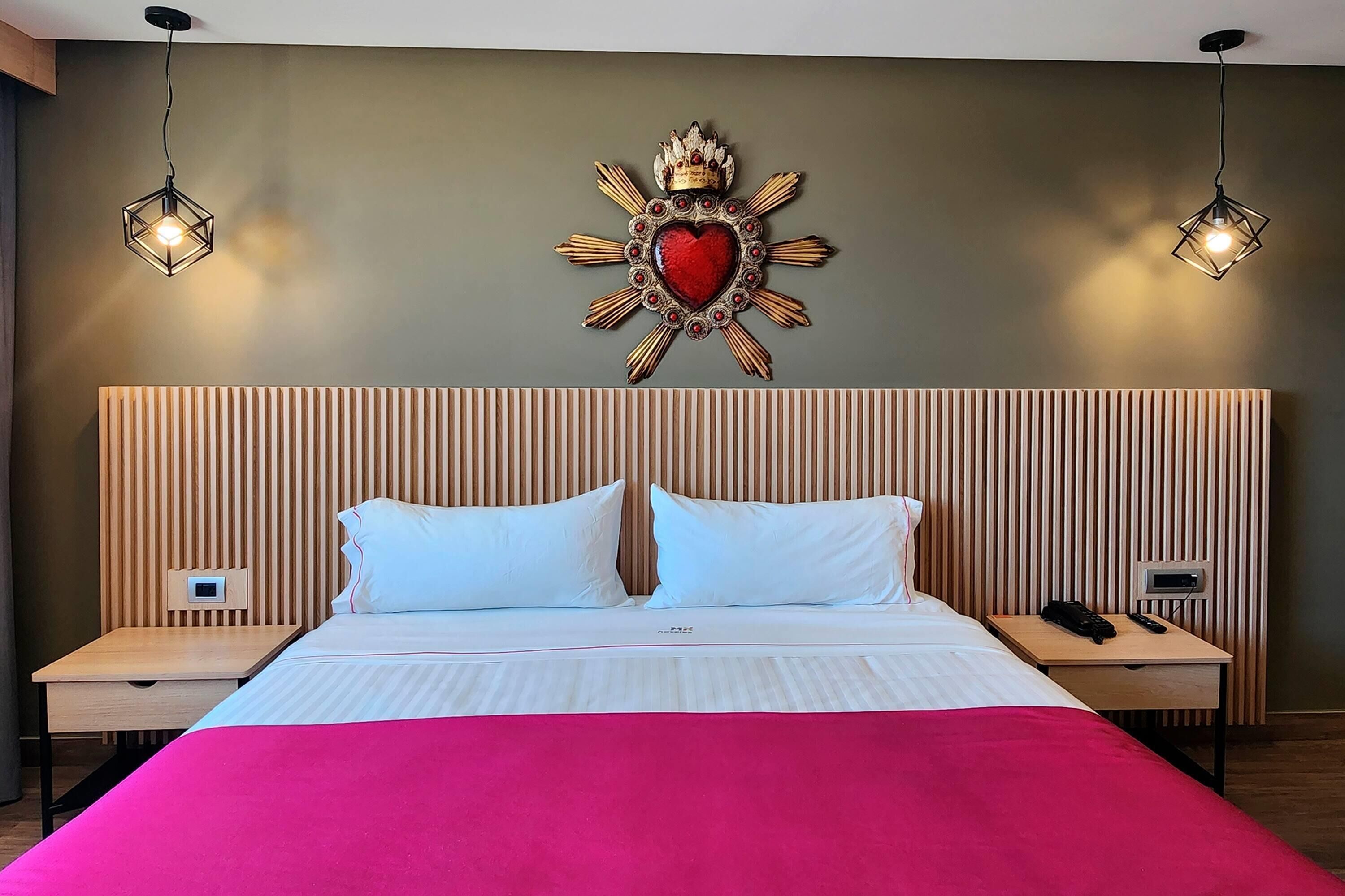 Quarto de hotel com cama, duas mesas de cabeceira, um objeto decorativo na parede e duas luminárias pendentes.