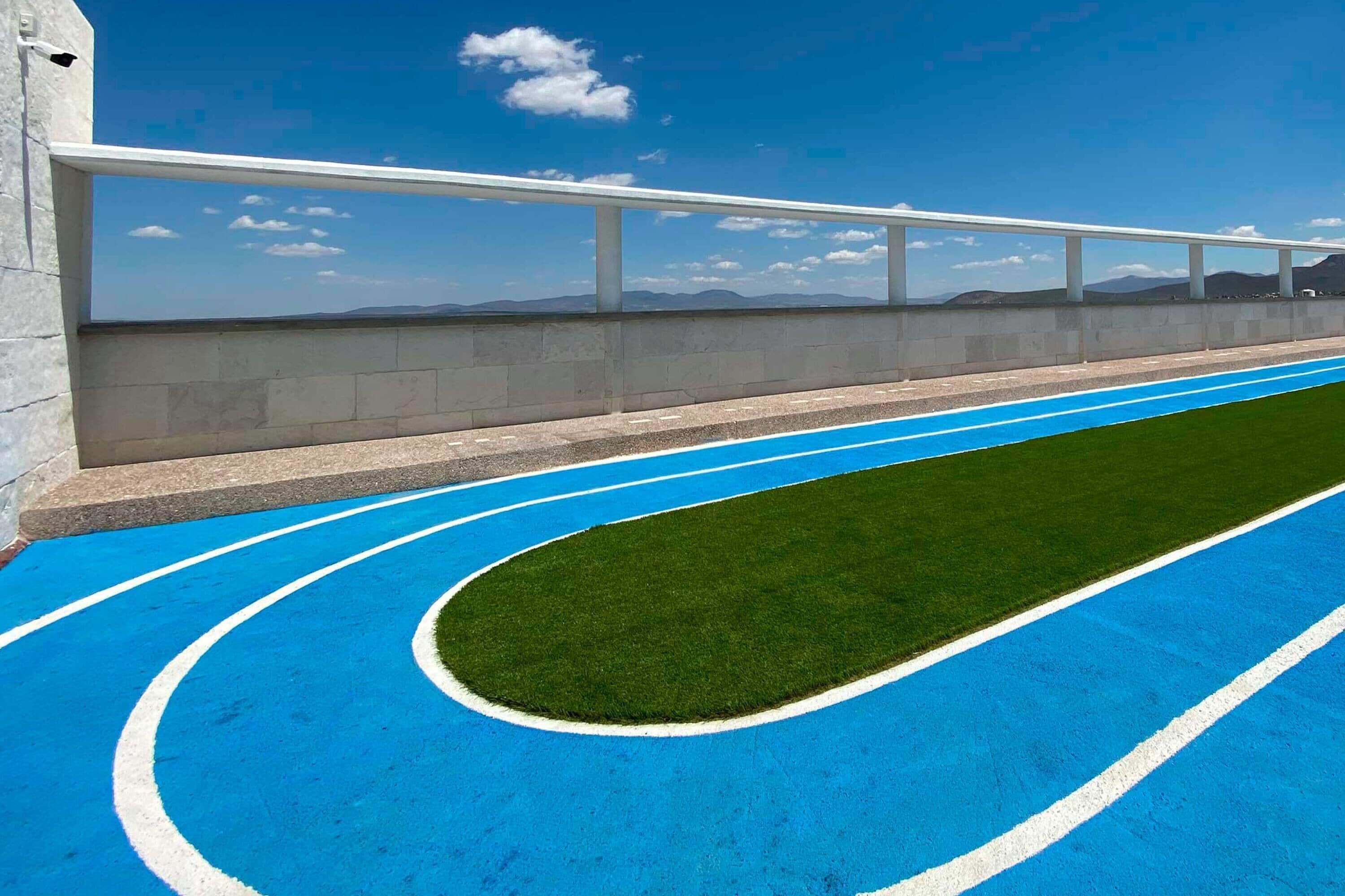Pista de atletismo azul con marcas blancas de carriles, rodeada por un muro de concreto y una barandilla blanca.