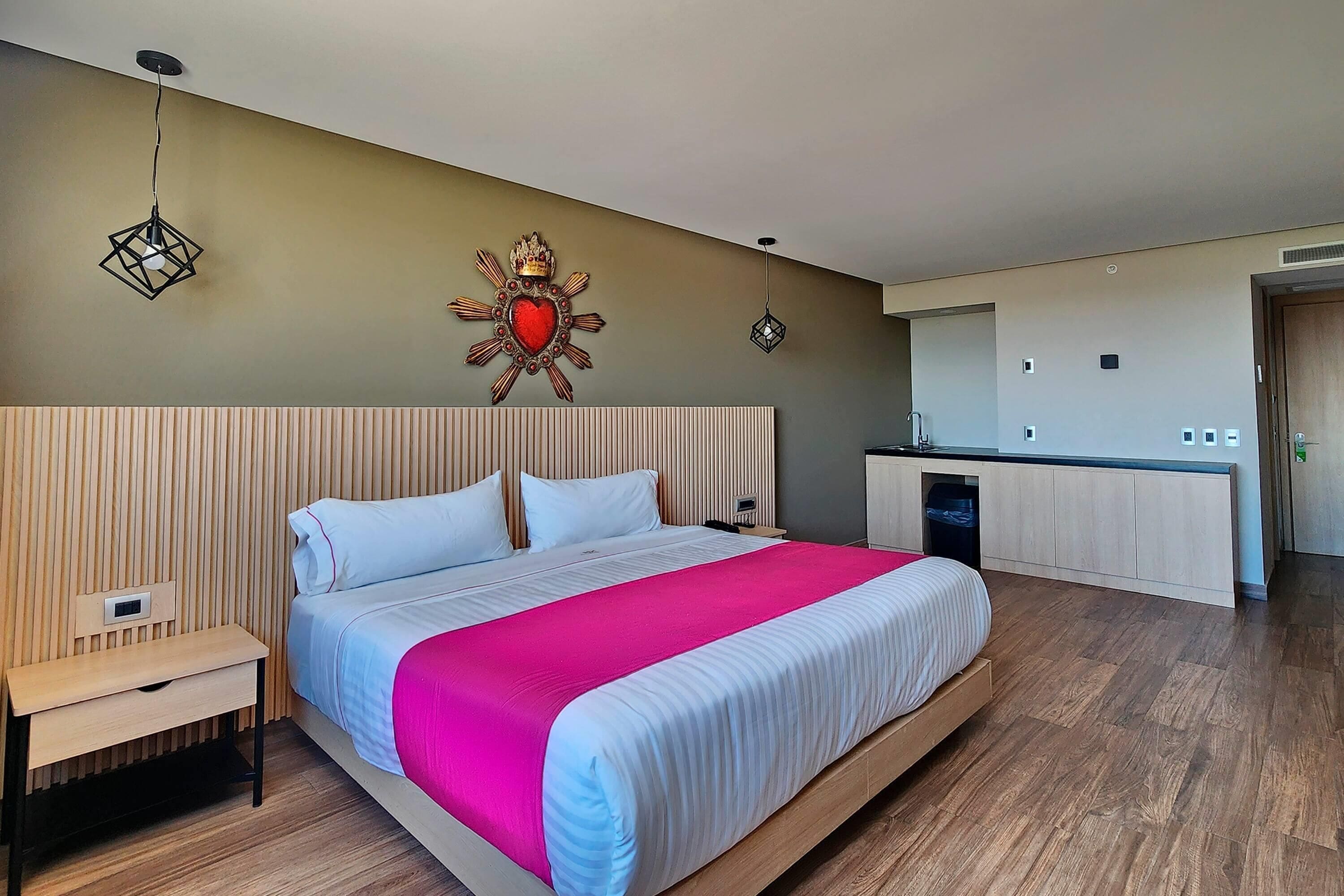 A modern hotel room with a large bed, a bedside table, a nightstand, a decorative wall piece, a hanging light, and a kitchenette area.