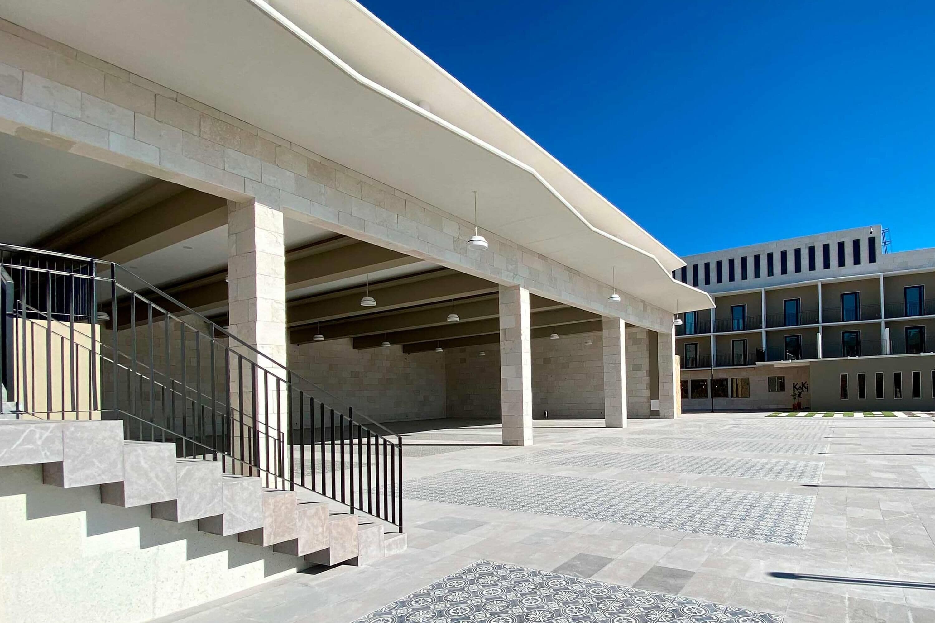 A spacious outdoor area with a modern architectural design, featuring a central staircase, multiple columns, and a patterned floor.