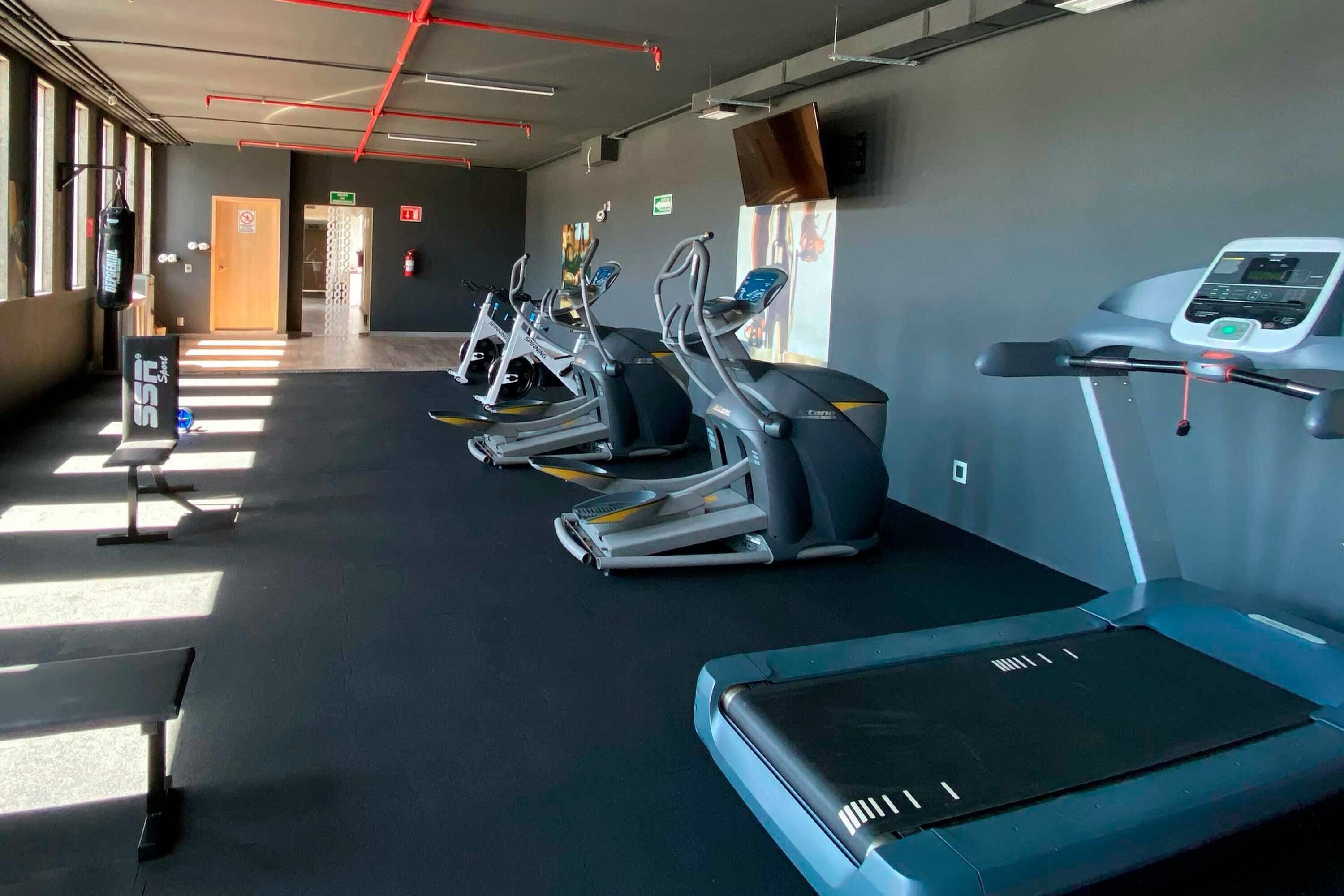 Un gimnasio con cintas de correr y elípticas, una banca y un televisor en la pared.
