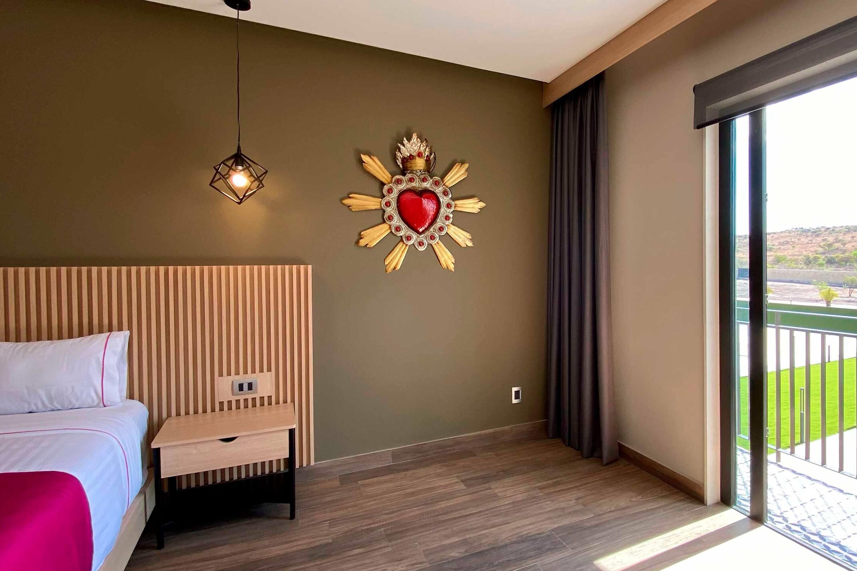A hotel room with a bed, a bedside table, a decorative wall piece, and a view of the outdoors.