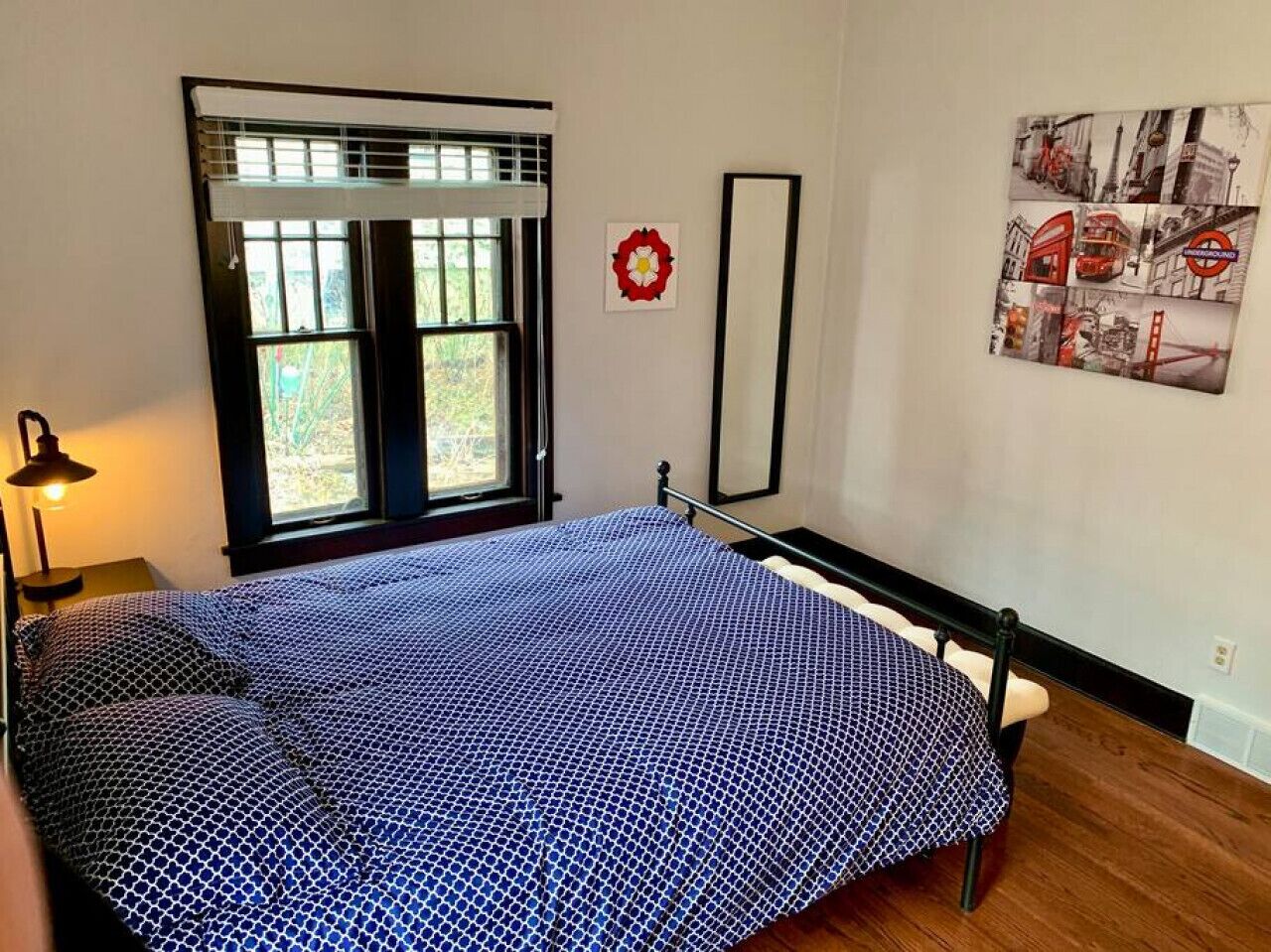 A bedroom with a bed, a window with blinds, a wall-mounted mirror, and a picture of London.