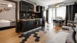 Une cuisine moderne avec des meubles de couleur noire, un plan de travail en bois et un sol carrelé à motifs hexagonaux. On y trouve un coin repas avec une table et des chaises, ainsi qu’un salon avec un canapé et un tapis à motifs.