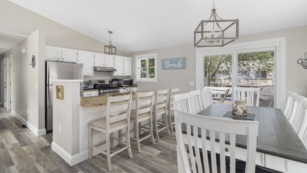 A kitchen with white cabinets, a stainless steel refrigerator, a wooden dining table with white chairs, and a chandelier.