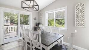 A dining area with a dark wooden table and white chairs, large windows with blinds, and a decorative wall piece.