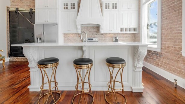 A kitchen with white cabinetry, a brick wall, and a large window. There are three stools with black seats and gold legs. - Historic downtown apartment with a view (Crowley)