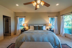 Une chambre avec un grand lit, un ventilateur de plafond, deux tables de chevet avec des lampes, une fenêtre avec des rideaux et une porte donnant sur un placard.