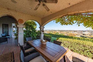 Une terrasse couverte avec une table en bois et des chaises en osier, un ventilateur de plafond et une vue sur un paysage lointain.