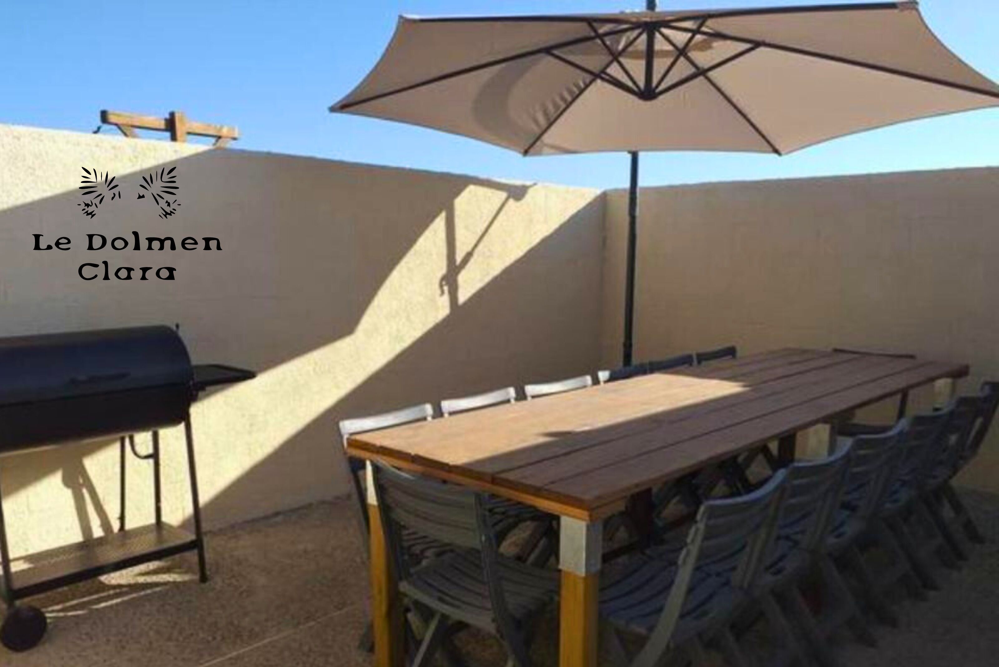 An outdoor patio with a wooden table and chairs, a barbecue grill, and a large umbrella providing shade.