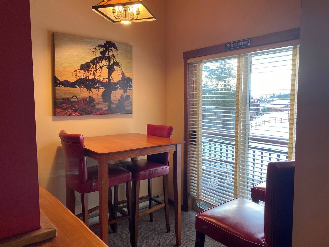 A corner room with a wooden table and two red chairs, a painting on the wall, and a window with blinds.