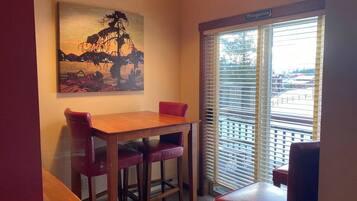 A corner room with a wooden table and two red chairs, a painting on the wall, and a window with blinds.