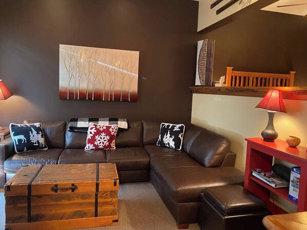 A modern living room with a sectional sofa, a wooden chest, and a red side table with a lamp.