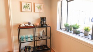 A kitchen area with a coffee maker, a shelf with glassware, and potted plants by a window.
