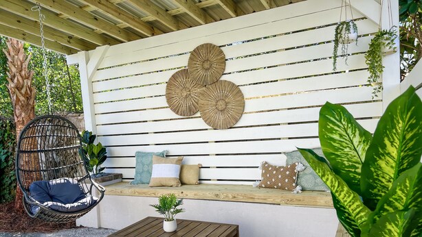 A wooden deck with a hanging chair, a bench with cushions, a small table, and large green plants.