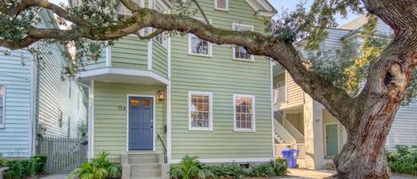 A two-story house with a green exterior, a front porch, and a large tree in front.
