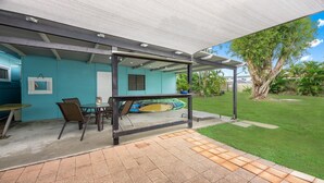 Un patio abrité avec une table et des chaises, une planche de surf, et une vue sur un jardin luxuriant et des arbres.