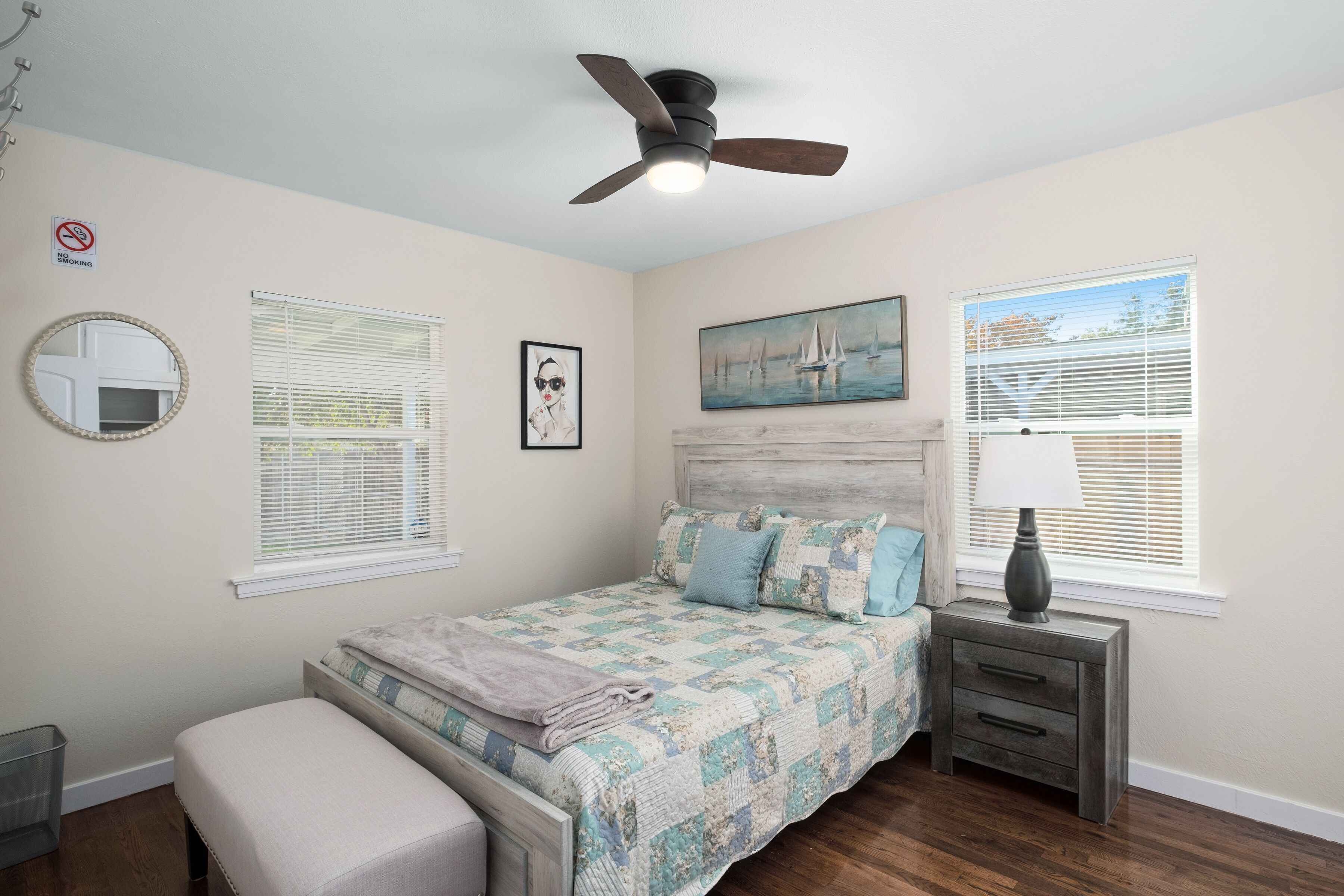 A bedroom with a bed, a nightstand, a ceiling fan, a window with blinds, a painting, and a mirror.