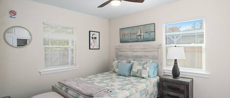 A bedroom with a bed, a nightstand, a ceiling fan, a window with blinds, a painting, and a mirror.
