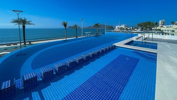 Ein Infinity-Pool auf dem Dach mit Blick auf den Ozean, umgeben von Palmen und modernen Gebäuden.