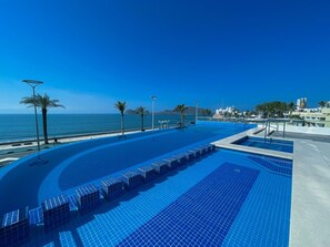 A rooftop infinity pool with a view of the ocean, surrounded by palm trees and modern buildings. - Varali Grand Hotel (Mazatlán)