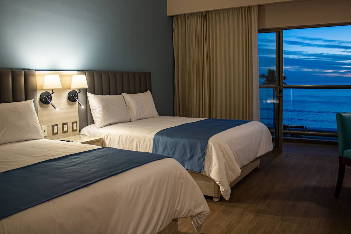 A hotel room with two beds, a large window with a view of the ocean, and a balcony with a chair.