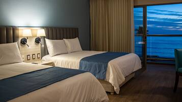 A hotel room with two beds, a large window with a view of the ocean, and a balcony with a chair.